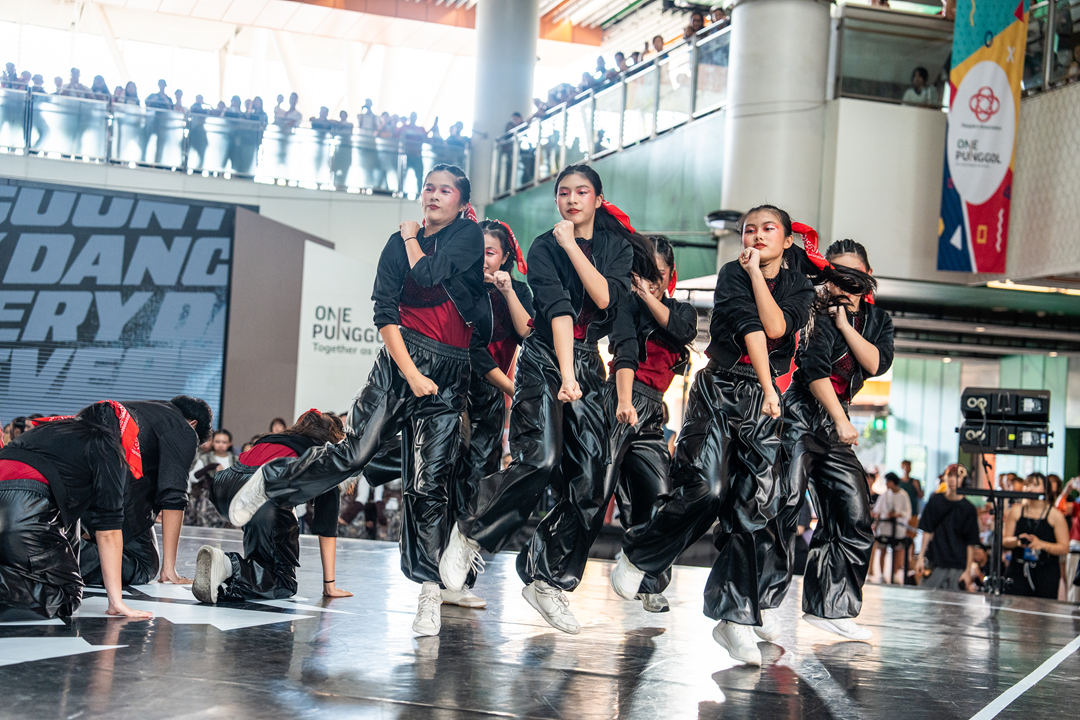 A dance crew wearing matching black outfits performs a dynamic synchronized dance routine on a lit stage inside a multi-level atrium.